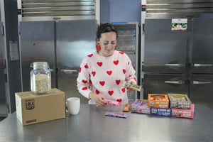 Mary in an industrial kitchen holding bars with boxes of bars on the table. Jar of oats on top of boxes to represent wholesomeness.
