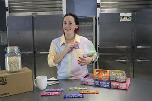 Mary in an industrial kitchen taking a bar out of her top shirt picket with bars and boxes of bars on the table. Jar of oats on top of boxes to represent wholesomeness.  Edit alt text